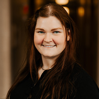 Corinne Sleeter stands in a low light hallway. She is wearing a black long sleeve dress shirt with a necklace. She has long brown hair, blue eyes and is smiling at the camera.