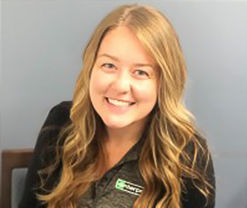 Julia Annarino sits in a wooden chair and looks up at the camera. She has long blond hair and is wearing a long sleeve black and gray Enterprise logo shirt.