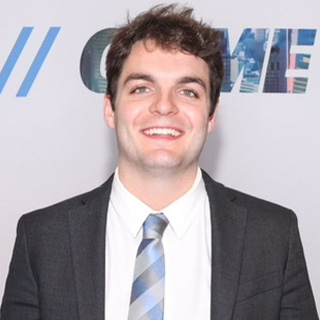 Carson Neuer stands in front of a branded step and repeat. He has messy dark brown hair, bushy eyebrows and a big smile. He is wearing a gray suit jacket, white button down and a blue and gray striped tie.