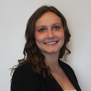 Kayli Kalish stands against a white wall and smiles. She has long brown hair that has been curled. She is wearing a white shirt and a black blazer with two silver necklaces.