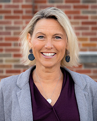 Lori Driessnack stands against a brick wall. She has chin length blond hair and smiles at the camera. She is wearing a light gray blazer over a wine color shirt. She has dark gray earrings and a silver necklace.
