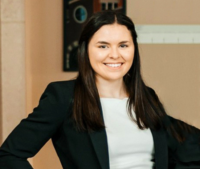 Lucy Forlastro wears a white shirt and black blazer. She has long dark hair. She has one hand on her right hip and is smiling at the camera.