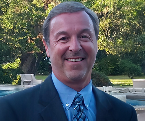 Lyndell Patrick smiles in an outdoor picture. He's wearing a blue suit jacket, blue button down and a patterned tie. He has short light brown and gray hair that is parted to the side and a short goatee..