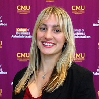 Mela Hammond poses in front of a maroon CMU step and repeat banner. She has long blond hair with choppy bangs and a nose ring. She is wearing a black blazer and a red shirt. She has on a silver necklace. Her lipstick is a bright red.