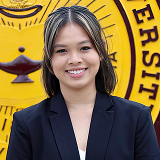 Mia Phan stands in front of the CMU seal on the Warriner mall. She has dark hair with blond highlights and is smiling. She has a dark suit jacket over a white shirt.
