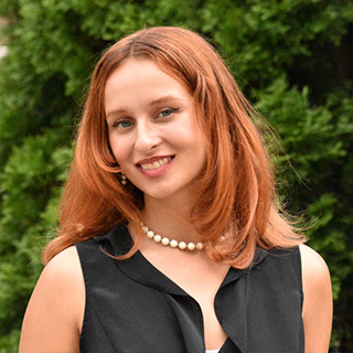 Reeve Katt has long red hair wears red lipstick while smiling at the camera. She is wearing a sleeveless black shirt with a deep neckline. She has on a pearl necklace. Behind her is green foliage.