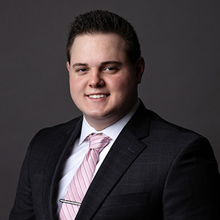Ryan Hammond wears a black suit jacket, white button down shirt and a pink tie with a silver tie bar. He has short dark hair with the front spiked up. He's smiling against a dark gray background.