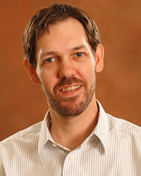 Sam Raisanen poses against an orange background and has dark brown hair and a short beard. He is wearing a button down white striped shirt. His ears are a noticeable feature.