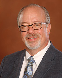 Steve Tracy poses against an orange backdrop. He has gray hair, with a top bald spot, and a gray goatee. He is wearing black framed glasses, a gray suit jacket, white button down and blue and silver tie.