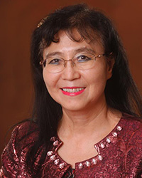 Vigdis Boasson poses against an orange backdrop. She has long black hair with short bangs. She has wire frame glasses and is smiling with red lipstick. Her shirt is maroon with white gemstone embellishments around the next