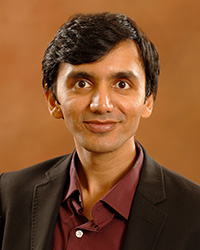 Vishal Shah poses against an orange background. He has black hair that sweep across his forehead. He is wearing a charcoal gray suit coat over a maroon button down shirt. The top button is undone.