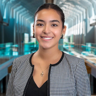 Yazmin Torres is standing in a large commercial atrium with large windows and blue glass. She wears a houndstooth black and white jacket with a black shirt, necklace and large black and white triangle earrings. She has dark hair that is pulled back.