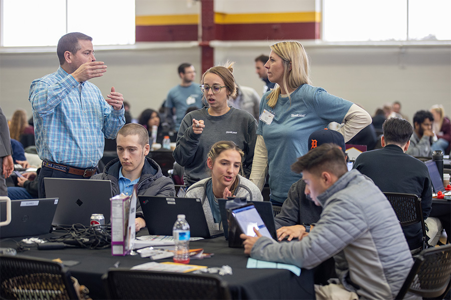 A group of students and advisors work together on laptops in a gymnasium.