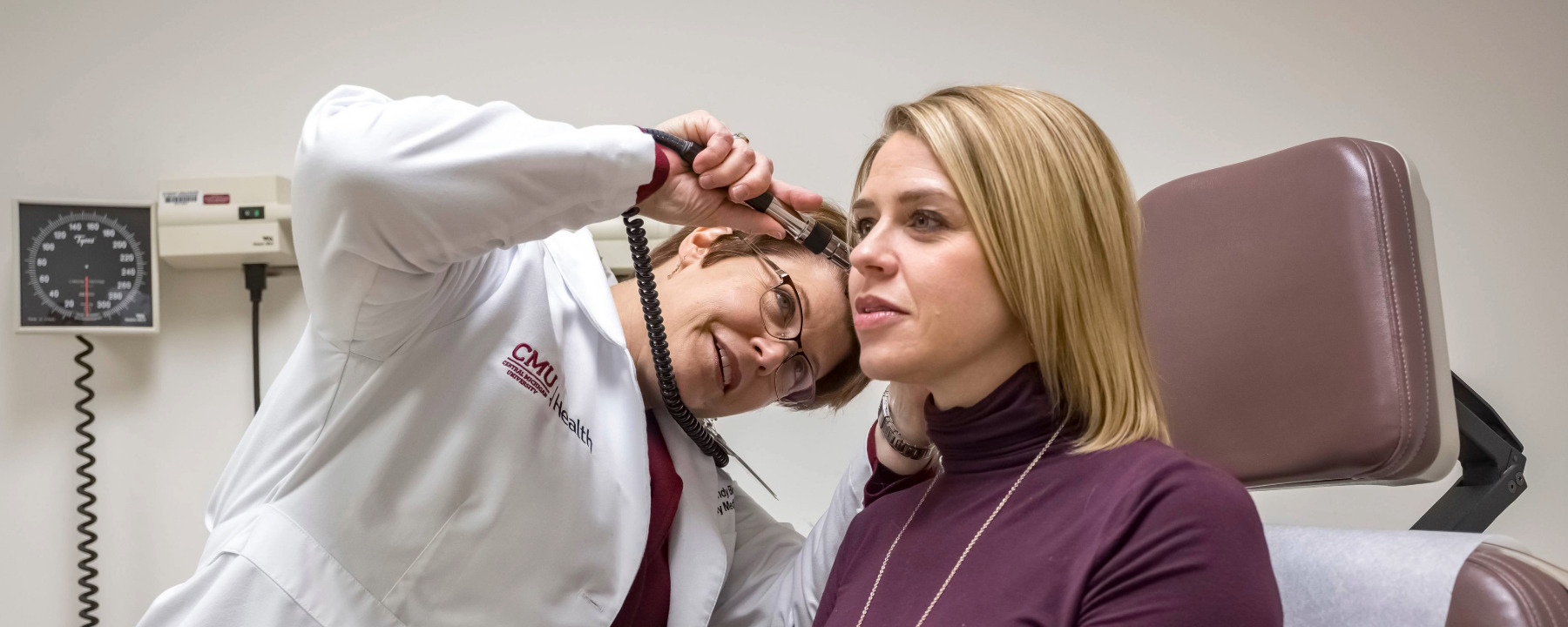 Wendy Biggs, M.D., in a medical exam room, using an otoscope to look in a female patient's ear.