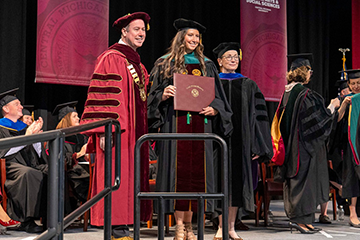 A medical school graduate in academic regalia smiles while holding her diploma on stage during a commencement ceremony with university leadership.