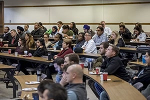 Medical students sit at tables in a lecture hall and look forward.