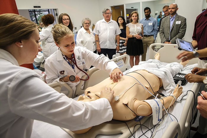 People observe as two young women in white lab coats inspect a patient simulator.