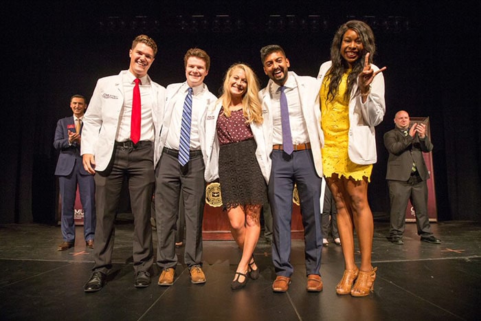 A group of students wearing white lab coats stand together and smile at a white coat ceremony.