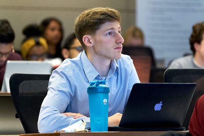 A young man with a black MacBook and a blue water bottle in front of him sits and looks straight ahead in class.