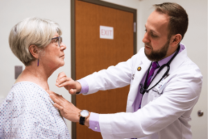 A medical student in a white coat examines an older patient wearing a hospital gown. The medical student is gently touching the patient's shoulder while speaking, demonstrating a calm interaction in a medical setting.