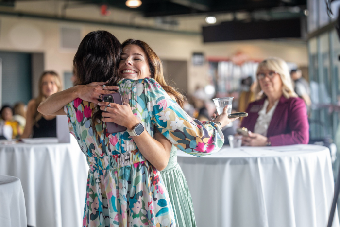 Match Day 2026 A medium shot shows two young women joyfully hugging at an indoor event. One, facing the camera, smiles and wears a colorful floral dress. The other, seen from behind, wears a matching dress. Other attendees are visible in the softly blurred background.