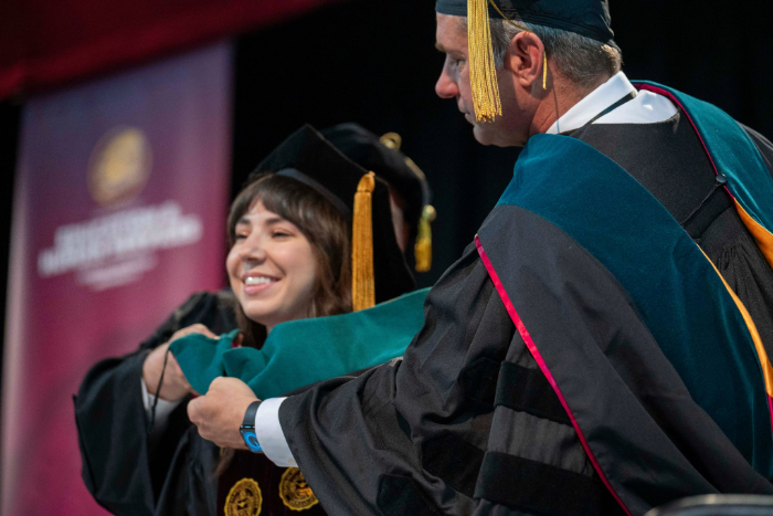 Student hooding Commencement 2026 A faculty member in academic regalia places a green doctoral hood over a smiling female graduate’s head during a hooding ceremony. Both wear black caps and gowns with gold tassels. A blurred maroon banner is in the background.