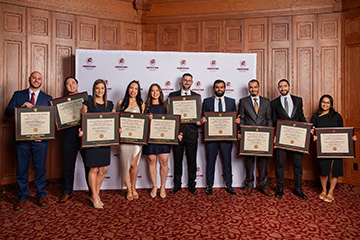 A group of 10 young men and women in formal dress hold their framed diplomas and smile at the camera.
