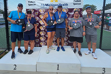 Three pairs of men and women holding pickleball paddles smile at the camera on a winner's podium.