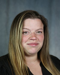 A woman with long blonde hair and a black shirt, smiles against a grey background.