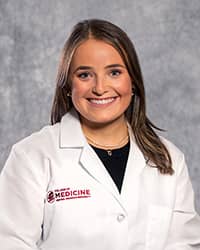 Blankenstein, Andie 200 x 250 web A woman with long brown hair wearing a black shirt and a medical white coat as she smiles at the camera.