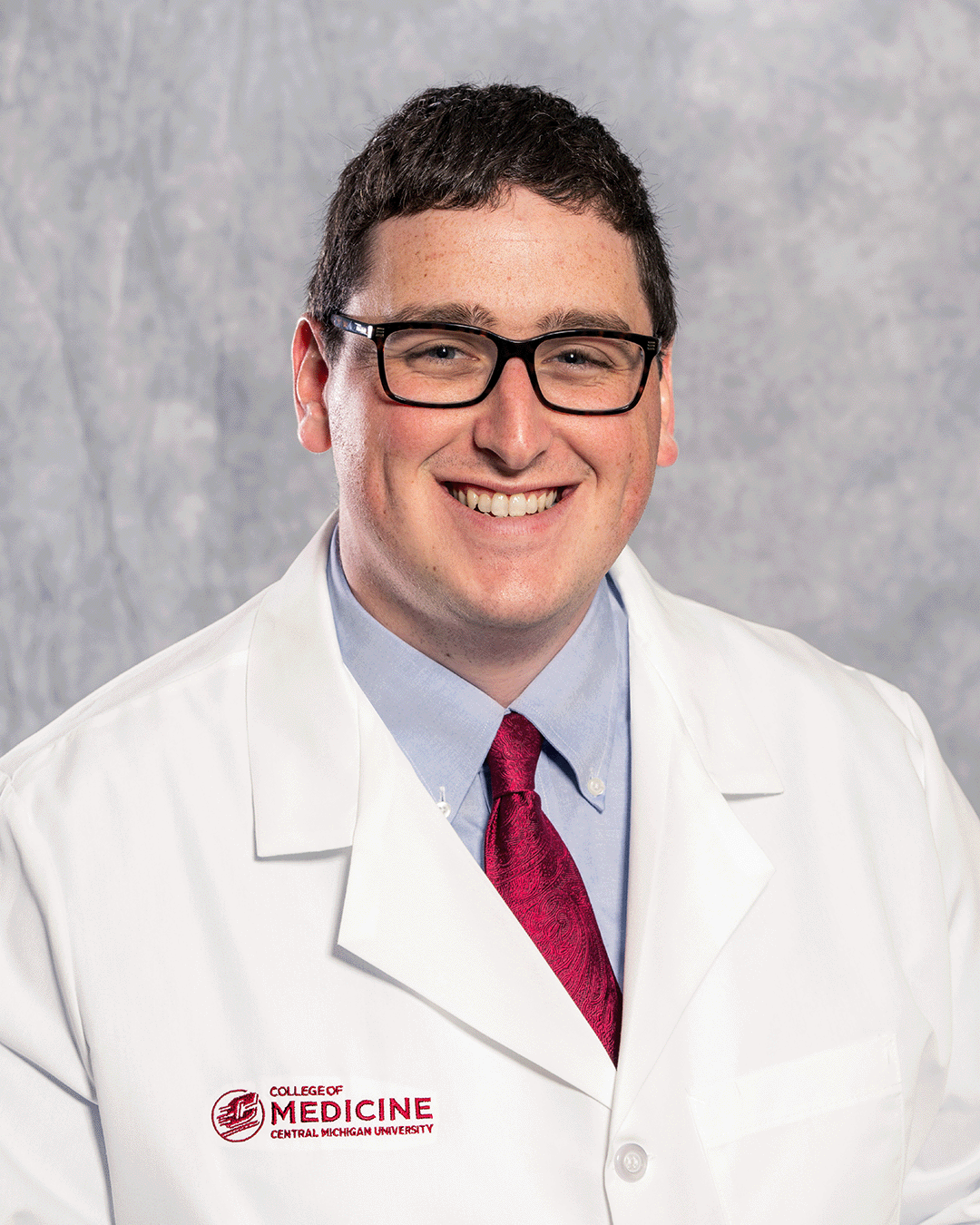 A male CMU College of Medicine medical student with rectangular black glasses is wearing his white coat as he smiles for the camera.