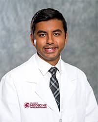 Gaur, Pranjal 200 x 250 A man with short, black hair, white collared shirt and dark plaid tie wears a medical white coat and smiles for a headshot.