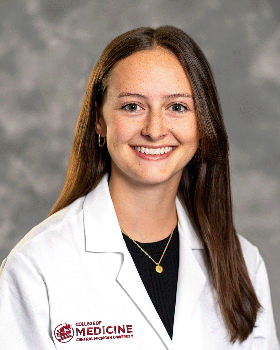 A female CMU College of Medicine medical student with long brown hair wearing her white coat over a black blouse as she smiles for the camera.