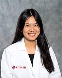 Huynh, Judy 200 x 250 A woman with long, black hair and a black blouse wears a medical white coat and smiles for a headshot.