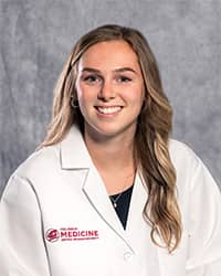 Manzo, Miranda 200 x 250 A woman with long, blond hair and a black blouse wears a medical white coat and smiles for a headshot.