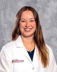 Miller, Madison 200 x 250 web A woman with long dark blond hair wearing a black shirt and a medical white coat as she smiles at the camera.