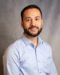 A man with short dark hair with a beard and mustache smiling wearing a light blue collared shirt.