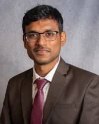 A man with dark hair and glasses wearing a light colored suit with a white collared shirt and pink tie.