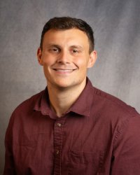 A man with short dark hair smiling wearing a maroon collared shirt.