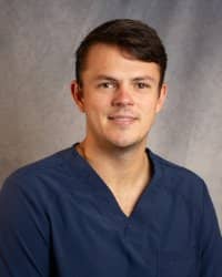 A man in a blue shirt smiles for a professional headshot in front of a grey background.
