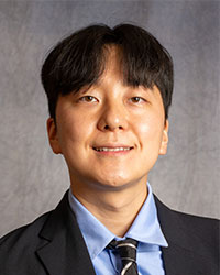 Jun Kim is wearing a black blazer, blue shirt, and black patterned tie.