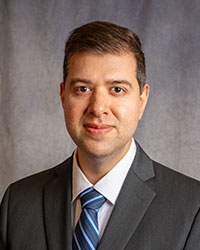 Azem Quereshi is wearing a grey blazer, white shirt, and blue striped tie.