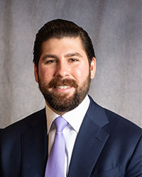 John Rahi is wearing a blue blazer, white shirt, and purple tie.