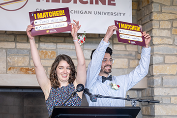 A woman and a man stand at a podium holding up signs and smiling at the camera.
