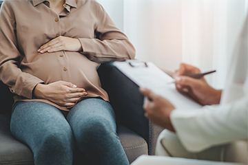 Pregnant person in beige shirt holds their belly while a doctor in a white coat takes notes on a clipboard.