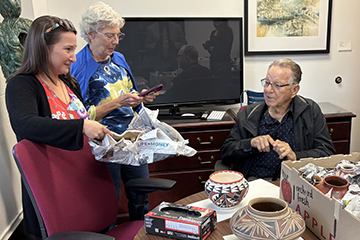 Megan Moreno unwrapping pottery from Jerry Thompson and his wife Mary. The three are gathered around a table that is scattered with more pottery.