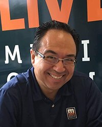 20230629_SCJMAlum_JohnGonzalez_001_200x250 Headshot of John Gonzalez in front of a banner for a television station.