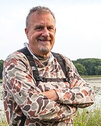 Headshot of Steve Jessmore. He is standing out in front of a lake with his camera equipment strapped onto his back.