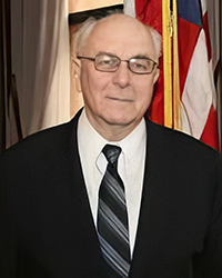 20250429_HallFame_FMMester_001_200x250 Headshot of Fred Mester in a suit and tie in front of an American flag.
