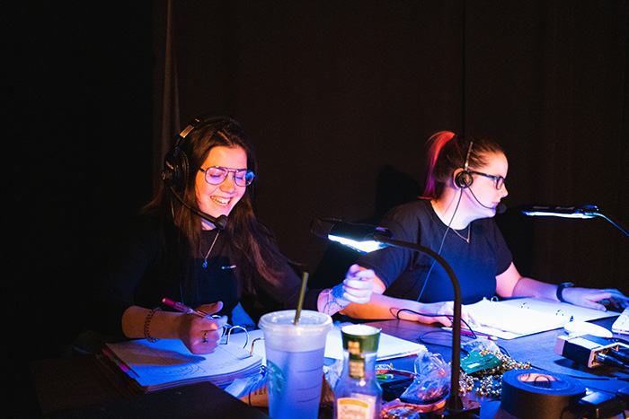 Two women sitting at a tech desk wearing tech headsets and looking at scripts.